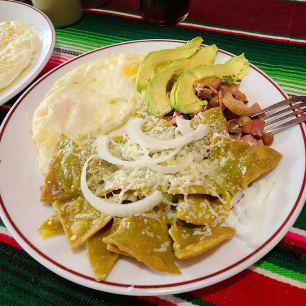 Chilaquiles verdes with fried eggs, avocado, beans, cheese, and onions on a white plate.