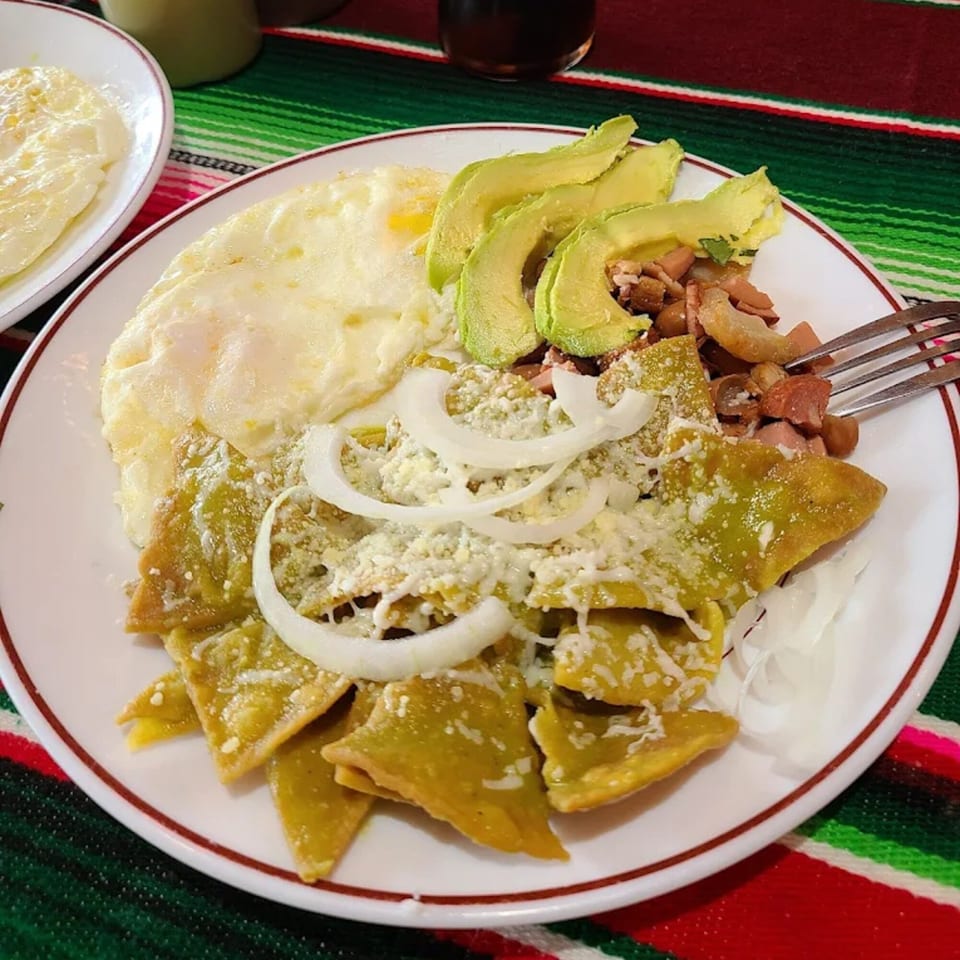 Chilaquiles verdes with fried eggs, avocado, beans, cheese, and onions on a white plate.