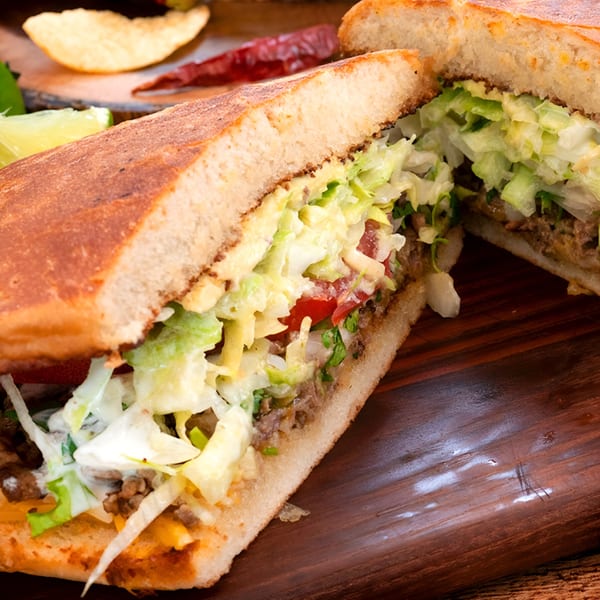 A Mexican torta sandwich, cut in half, filled with meat, lettuce, tomatoes, and sauce.