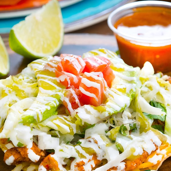 A Mexican tostada topped with chicken, lettuce, tomatoes, and sour cream, with lime and salsa.