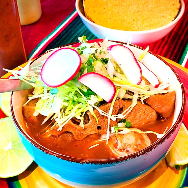 A vibrant bowl of Mexican menudo soup, garnished with radishes, cabbage, and lime wedges.
