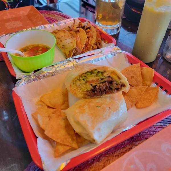 Mexican food spread with a burrito, tacos, chips, soup, and drinks on a table.