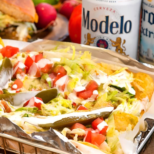 Crispy tacos topped with lettuce, tomatoes, sour cream, and pickled jalapeños, with Modelo beer cans.