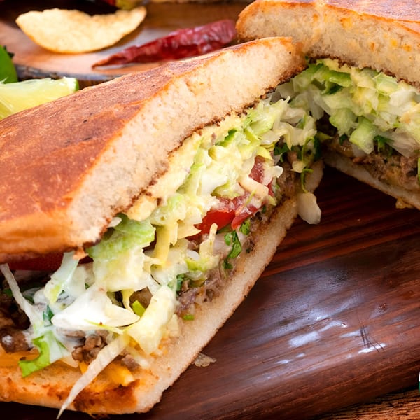 A large Mexican torta sandwich, cut in half, filled with meat, lettuce, tomato, and sauce.