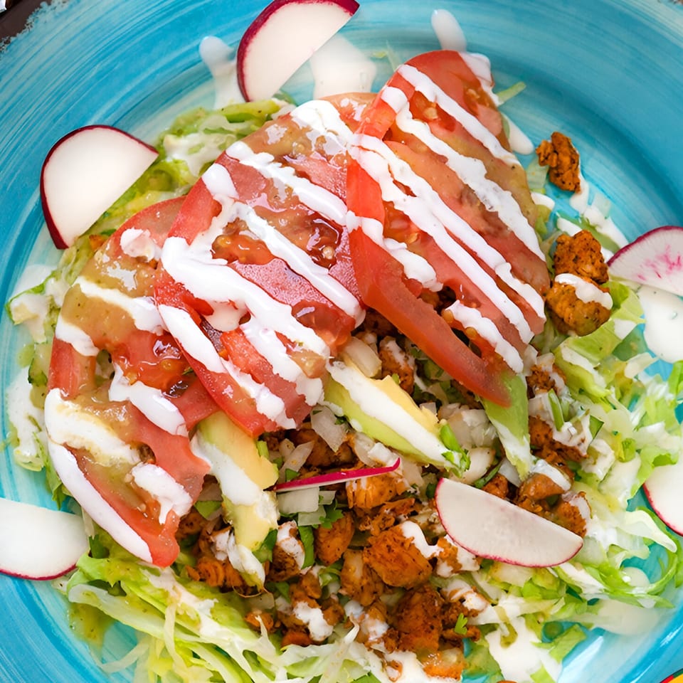 Mexican salad with chicken, lettuce, tomatoes, avocado, radishes, and crema on a blue plate.