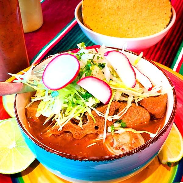A vibrant bowl of Mexican menudo soup, garnished with cabbage, radishes, and lime slices.
